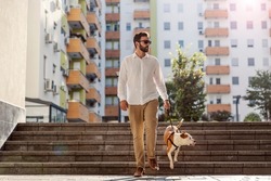 A busy man dressed smart casual, with sunglasses is walking his dog in the urban exterior. The dog descending stairs. A man with a dog in a walk in the urban exterior.