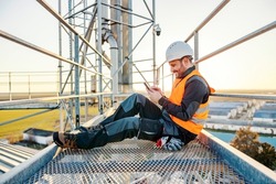 An industry worker sitting on metal construction on a break and scrolling on the phone.