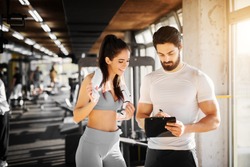 Young slim fitness girl standing with a towel near handsome trainer while showing her schedule for next week in the gym.