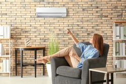 Young woman switching on air conditioner while sitting in armchair at home