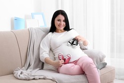 Beautiful young pregnant woman holding baby shoes and sitting on sofa at home