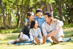 asian family with two children having fun sitting on grass talking chatting outdoors in park