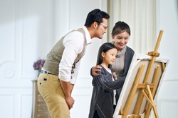 beautiful little asian girl with long black hair making a painting on canvas while parents standing behind watching.