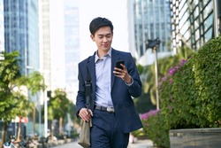 young asian businessman looking at messages on cellphone while walking in the street in downtown of modern city