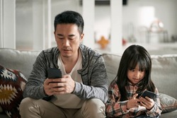 young asian father and seven-year-old daughter sitting on family couch using cellphone together looking serious