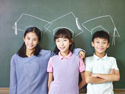 three asian elementary school children standing underneath chalk-drawn doctoral hats in front of blackboard.