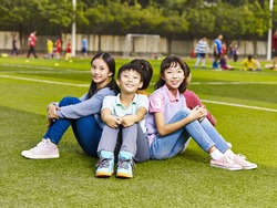 group of happy smiling elementary school boys and girls sitting on grass of playground.