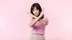 portrait young serious asian woman with cross arm gesture showing stop, no, wrong, denial, rejection sign isolated on pink pastel studio background. deny and negative expression symbol concept.