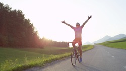 LENS FLARE: Thrilled bicycle rider celebrates win in bike race across countryside by outstretching arms and looking into the golden evening sky. Pro road cyclist rejoicing after completing workout.