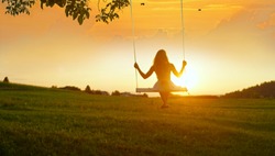GOLDEN LIGHT SILHOUETTE: Unrecognizable girl in white dress swaying on a tree swing on peaceful evening. Lady sitting on a wooden swing and looking at golden sunset. Young woman swinging at sunrise