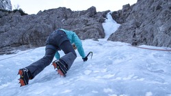 BOTTOM UP, CLOSE UP: Caucasian woman ice climbing in the Julian Alps stops and looks at her crampons Fearless female tourist goes ice climbing up the gorgeous icy stream running down the rocky cliff