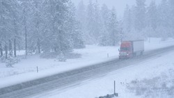 Red semi truck hauls a heavy cargo container across the state of Washington and through a snowstorm. Freight lorry navigates the slippery country road in the low visibility during an intense blizzard.