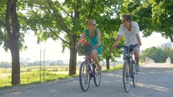Young Caucasian couple smiles while riding bicycles down a scenic green avenue. Beautiful blonde girl and her handsome boyfriend riding their bikes along a picturesque sunlit park in the summertime.