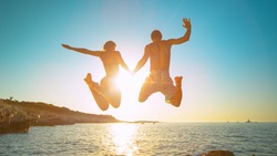 LENS FLARE: Carefree tourists hold hands while jumping into the refreshing sea at golden sunset. Young and energetic traveler couple holds hands while diving into the sea on a sunny summer evening.