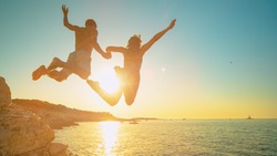 LENS FLARE: Carefree tourists hold hands while jumping into sea at sunset. Cheerful young woman holds her athletic boyfriend's hand as they jump off a cliff and into the cool sea at golden sunset.
