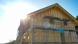 CLOSE UP, LENS FLARE: Bright spring sunbeams shine on a large wooden house being built in countryside. Scenic view of an unfinished cross-laminated timber house being built in rural area on sunny day.