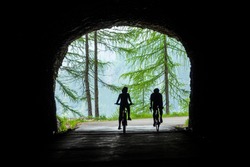 Two unrecognizable active tourists ride a modern electric and road bicycle out of an old dark tunnel as they explore the scenic mountain roads leading across the picturesque Julian Alps in Slovenia.