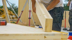 CLOSE UP, DOF: Unrecognizable male builder picks up a CLT beam from a workbench at an unfinished housing project. Contractor carries a glued laminated board across the bustling construction site