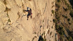 AERIAL: Young male rock climber with rope ascending up the sunlit limestone wall. Man climbing up the wall and searching for good grips. Adrenaline outdoor activity in beautiful natural environment.