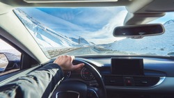 view from luxury car inside with part of interior gps screen with driver male hand on  the steering wheel during bright snowy sunny day on straight ice road with snowy mountains in background 