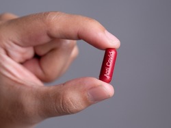 Male hand holding a medical red capsule pill between fingers against gray background,Coronavirus,Covid-19 Cure,Anti-Covid 19,anti flu drug pill