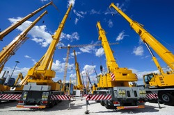 Mobile construction cranes with yellow telescopic arms and big tower cranes in sunny day with white clouds and deep blue sky on background, heavy industry 