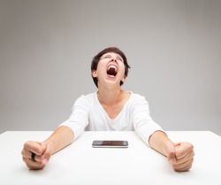 Frustrated woman throwing a temper tantrum clenching her fists and screaming at a mobile phone on the table in front of her