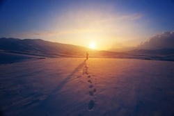 Lonely woman standing far away in bright yellow sunset light, traveling with backpack in the mountains covered with snow