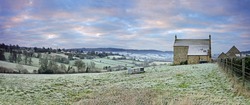 A panorama view of a cold and frosty sunrise over an isolated farm house and a distant view of the Cotswold village of Painswick, Gloucestershire, England, UK