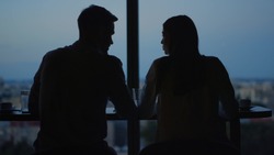 Business people silhouettes talking at workplace in dusk office. Back view of young couple flirting in office on evening city background. Love couple at dating in front of panoramic windows