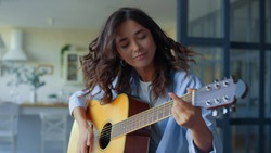 Excited girl playing acoustic guitar in living room. Happy musician playing chords on string instrument. Positive guitarist moving head during song. Musician performing musical composition on guitar