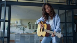 Smiling girl performing musical composition on acoustic guitar at home. Positive musician playing song on string instrument. Female guitarist playing on musical instrument in living room