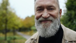 Close up portrait of bearded man looking at camera outdoors. Happy smiling senior enjoying relaxed atmosphere on street. Cheerful elder man posing for camera outside with autumn background.