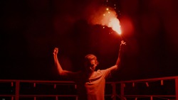 Aggressive man burning signal flare on night roof. Serious football fan standing with red fire outdoors. Man protest concept. Male protester with light at urban background.
