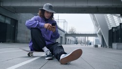 Young sporty man reading message online on phone on skateboard. Stylish hipster sitting on skate with mobile phone outdoor. Cool skater guy holding smartphone outside.