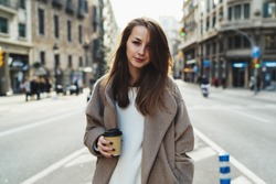 Half length portrait of a smiling hipster girl dressed in trendy street outfit holding take away coffee cup in hand and looking at camera while strolling outdoors. Student girl walking down the street