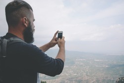 Young hipster is taking photo by smart phone on the peak of mountain