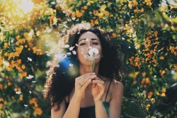 Young model look caucasian female with long dark curly hair wearing a black summer dress is blowing the dandelion at the camera while sitting on a blurred flowers background. Flare light.