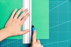 Female young hands cutting a green sheet of paper with a cutter and a metal ruler on a cutting board. Flat lay image with copy space.