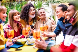 Friends toasting drinks at cocktail bar with face mask - New normal life concept with happy people having fun together drinking spritz at restaurant - Vivid filter with focus on black hair woman