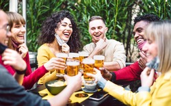 Multicultural people toasting beer wearing open face mask - New normal life style concept with friends having fun together at brewery bar garden - Warm filter with focus on woman in yellow clothes