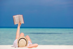 Young girl is reading a book lying on tropical white beach