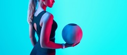Portrait of a fit woman standing holding a medicine ball. Copy Free space. Slim caucasian cross fit woman with fitness ball on bright neon cyan background