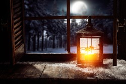 Dark wooden old winter window sill cover of snow and frost.Retro big lamp with candel and orange light.Landscape of winter forest and cold december night. 