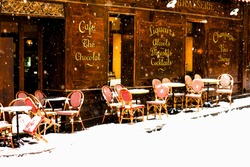 Classic old street brasserie in Paris viewed under the snowfall. Terrace circular tables and woven chairs are covered with fresh snow. Parisian cafe under the snowfall.