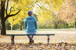 Depressed and sad old woman on bench in autumn park, from back angle