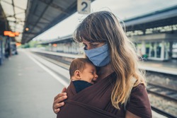 A young mother with her baby in a sling is wearing a face mask at a train station