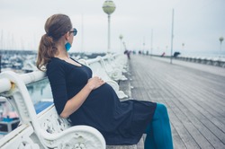 A young pregnant woman is sitting on a bench on the pier