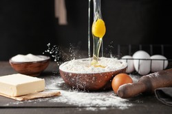 The raw egg is poured into a bowl of flour. Chef hands pouring flour powder on raw dough using sieve on a black background, Cooking process. 