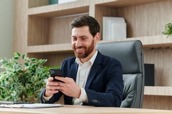 Businessman is smiling and typing a message on his smart phone in office. He is in a suit, workplace in modern and nice office